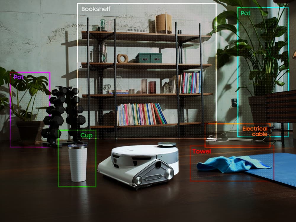 Robot aspirador Samsung Bespoke Jet Bot AI+ en un salón, con recuadros que marcan los objetos que reconoce: estantería, macetas, taza, toalla y un cable eléctrico